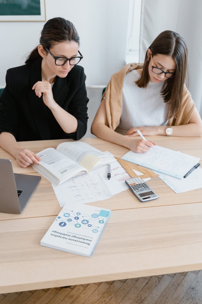 Two women engaged in focused study with books and laptops at a wooden desk indoors.