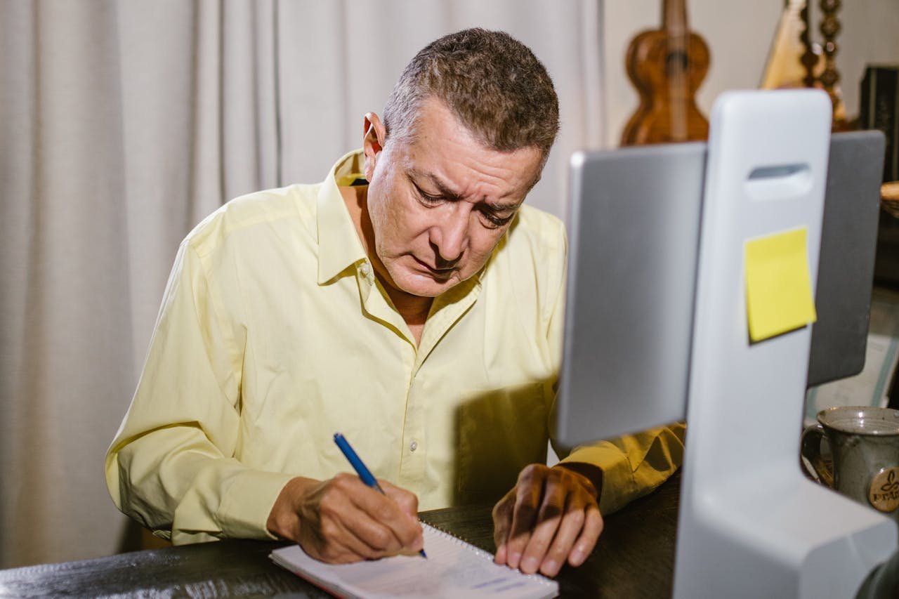 Elderly man focused on writing at home, sitting at a desk with a digital tablet.